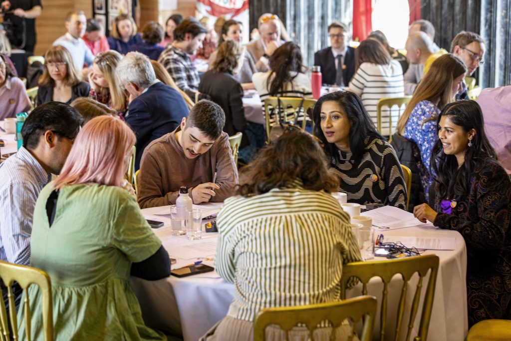 Group of people at a conference, with a man in the picture writing and people talking to each other on a roundtable
