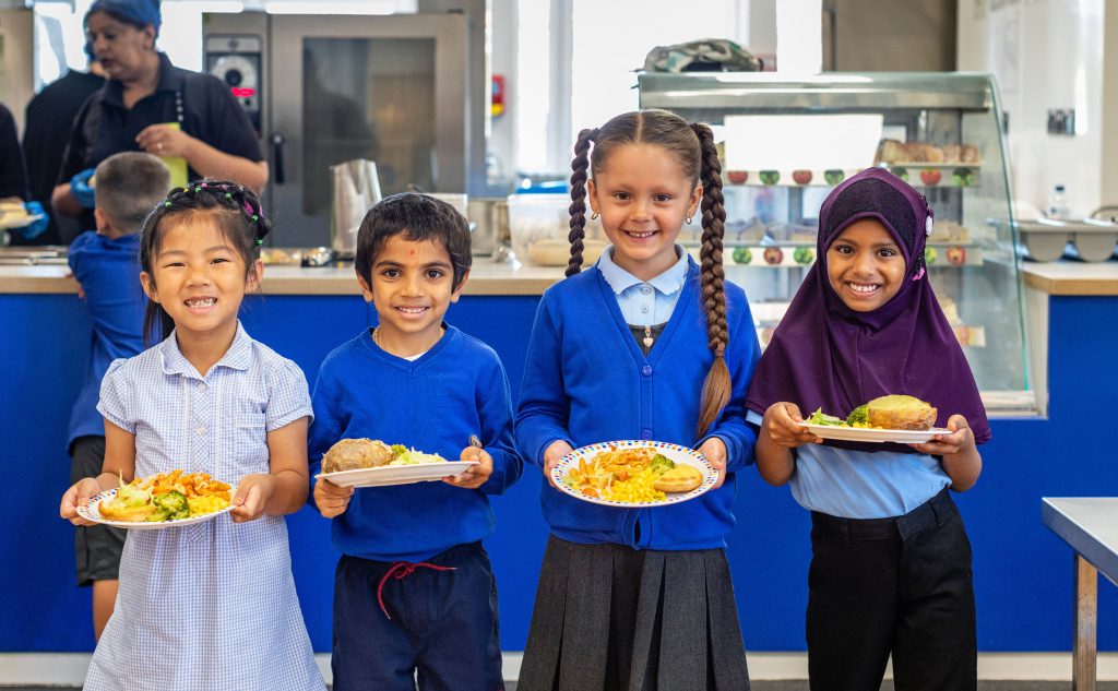 Primary school children holding school plates of food, stood in a line facing the camera, smiling