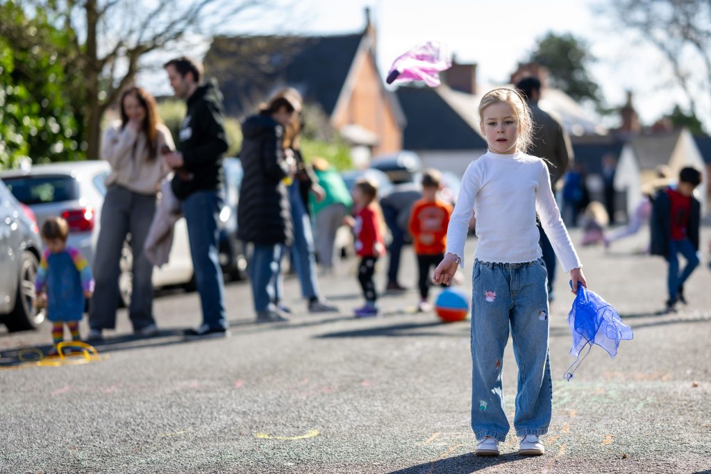 A girl playing in a street, with families and children blurred in the background