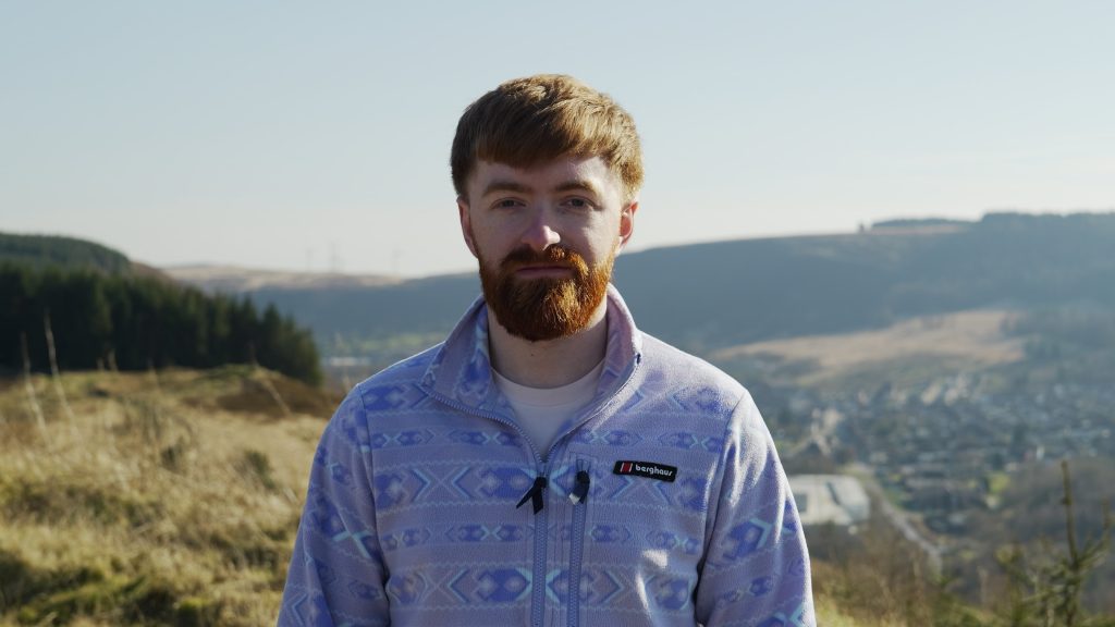 Dyn ifanc yn edrych ar gamera ar ben mynydd ar ddiwrnod cymylog