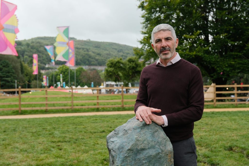 Safodd Derek Walker, Comisiynydd Cenedlaethau'r Dyfodol Cymru, y tu allan mewn cae gyda'i ddwylo wedi'u gosod ar ben craig, yn wynebu ymlaen.